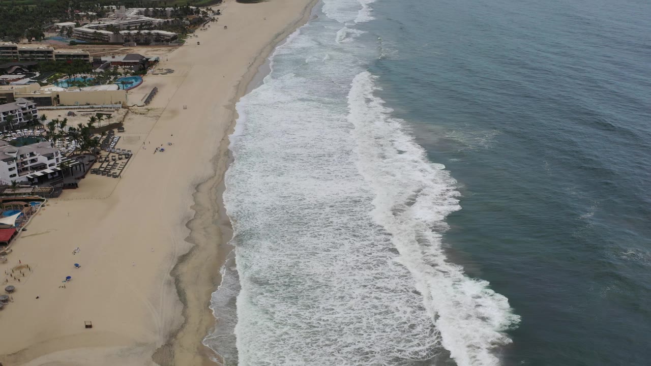 Los Cabos shoreline from above, with foamy waves filling the center, open ocean to the right, and low beachfront buildings with blue pools along the sand.