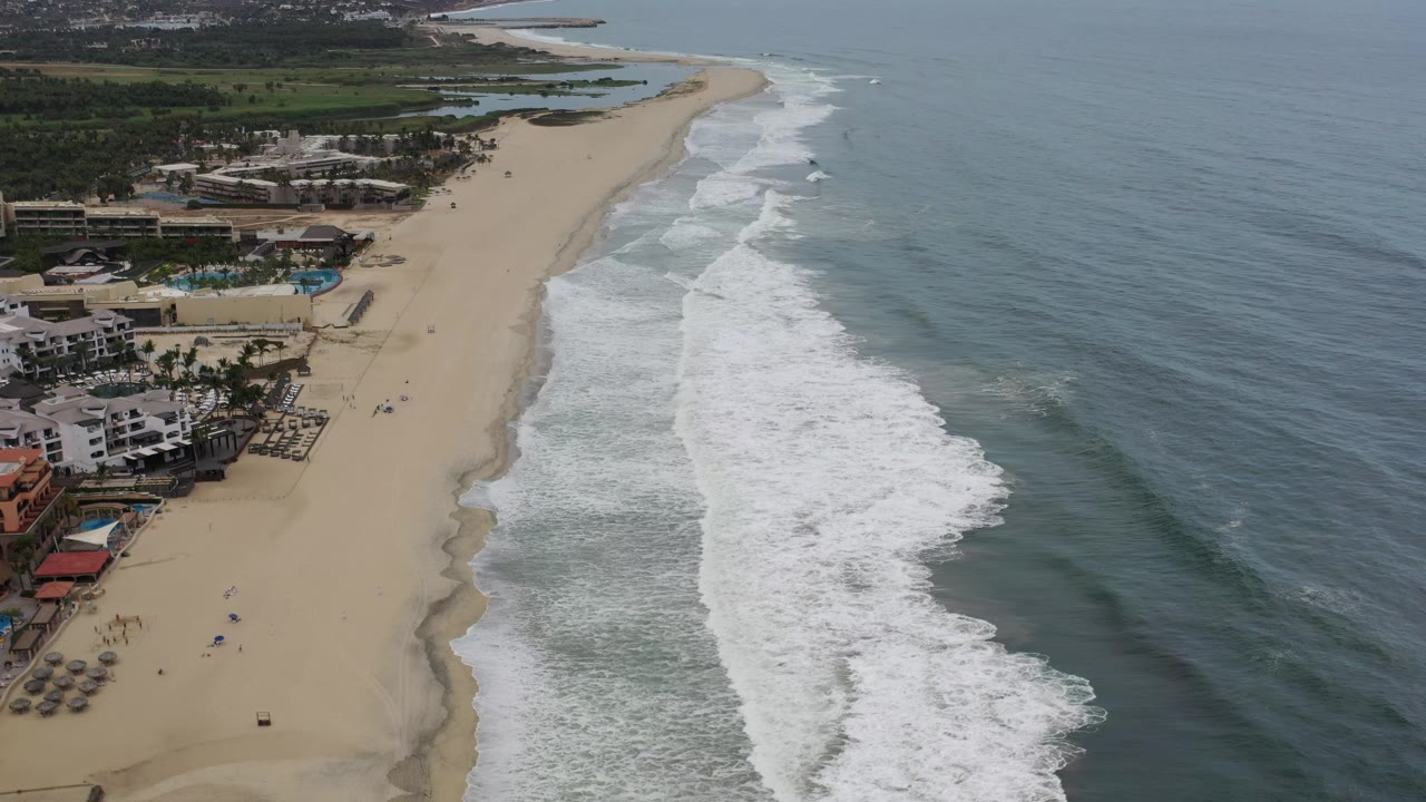 Elevated Los Cabos coastal frame showing waves spreading farther up the sand, resort-like buildings near the lower left, blue-gray ocean to the right, and no visible people.