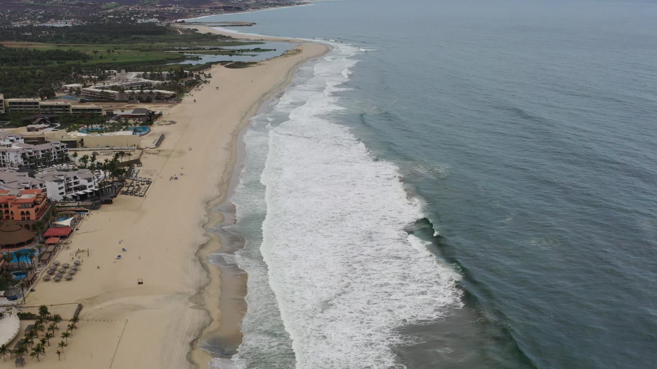 Aerial view of sandy beach, surf, ocean, beachfront buildings, and distant hills.