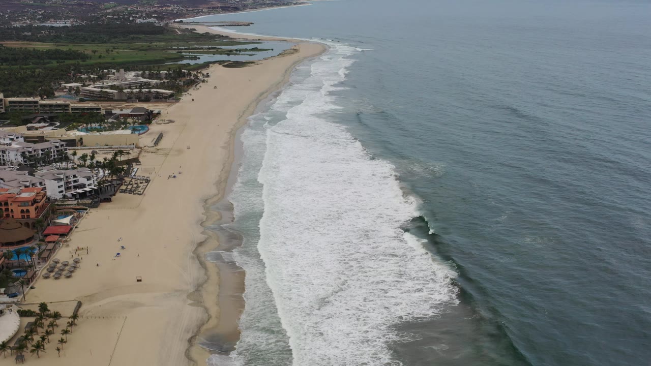 High aerial frame in Los Cabos showing the coastline stretching into the distance, a cluster of beachfront buildings at lower left, foamy waves along the beach, open ocean at right, and no visible people.