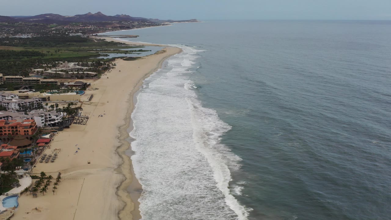 Opening frame of a Los Cabos, Mexico coastline with low buildings at lower left, a long sandy beach, white surf bands, blue-gray ocean, distant hills, and no visible people.