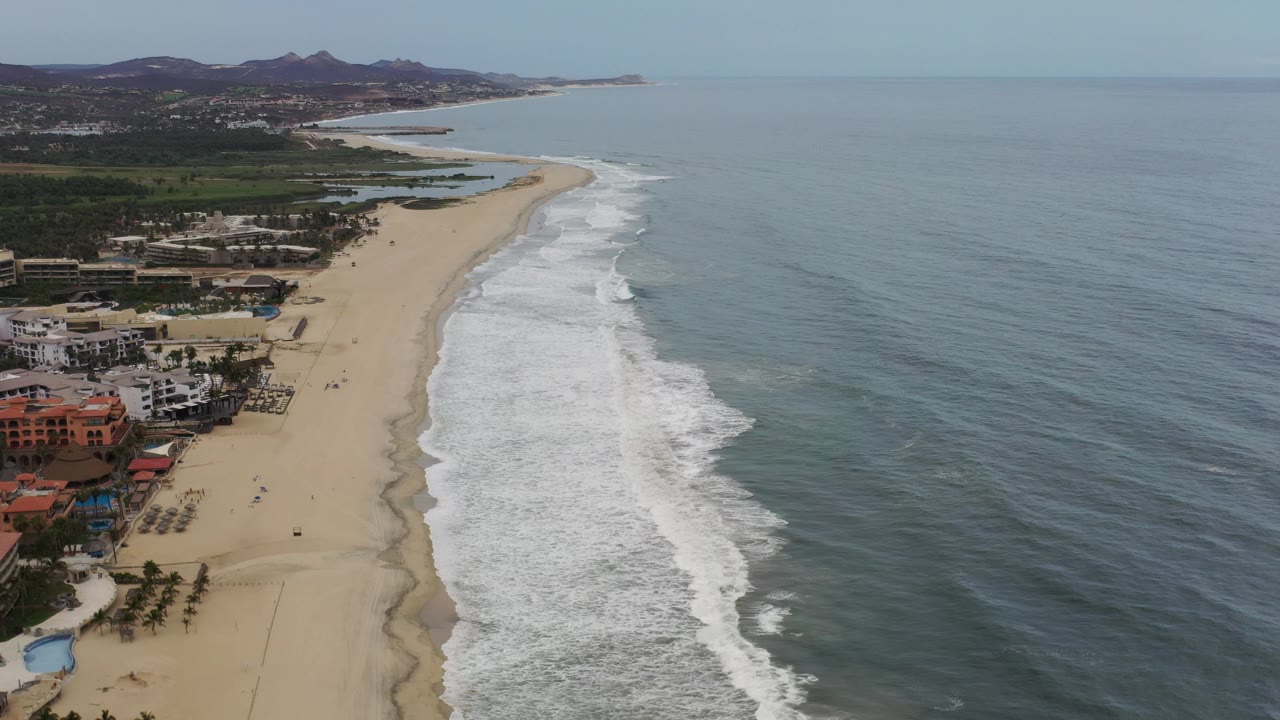Wide drone-like view of beach, waves, ocean, inland greenery, and hills.
