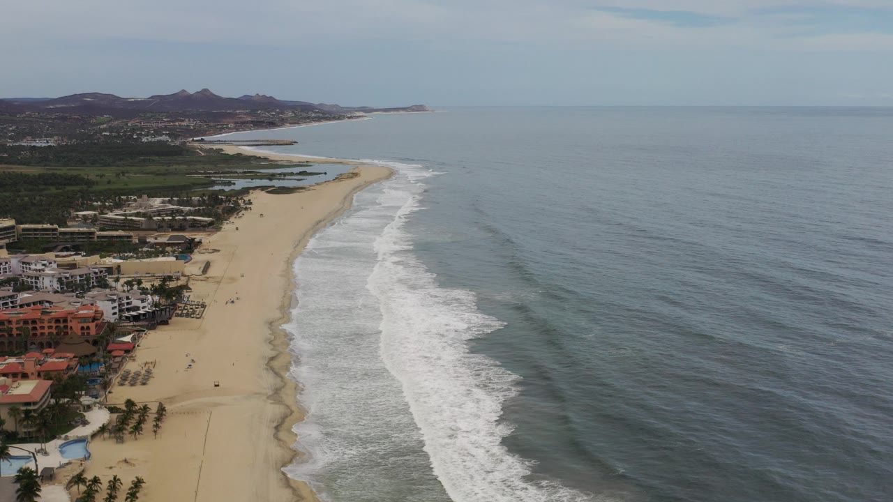 Opening aerial view over Los Cabos, Mexico with beachfront houses at left, a long sandy beach, white surf bands, gray-blue ocean, distant hills, pale sky, and no visible people.