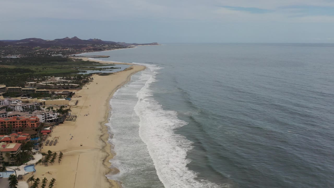 Wide coastline aerial with houses, beach, surf, and ocean.