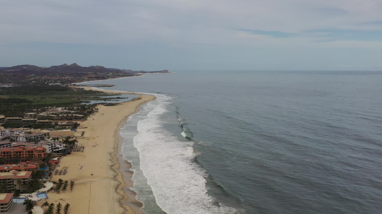 Aerial view of a sandy beach beside ocean surf and resort buildings.