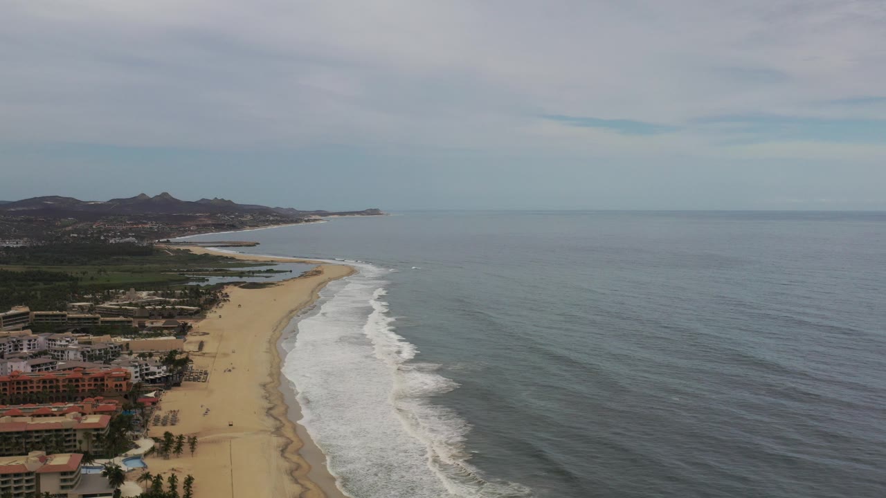 Opening frame of a Los Cabos, Mexico coastline from above, with beachfront buildings at lower left, white surf on tan sand, gray-blue ocean to the right, distant hills on the horizon, and no visible people.