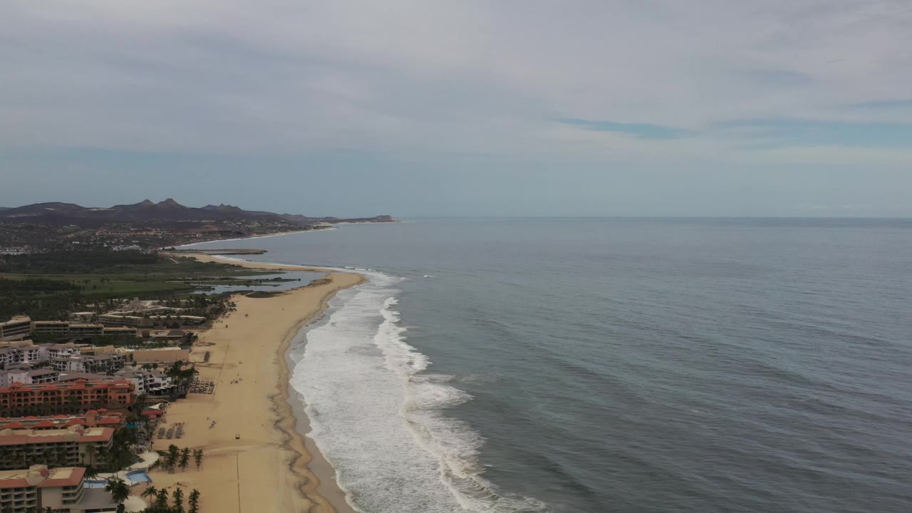 Los Cabos aerial frame with the shoreline slightly farther along the curve, beachfront buildings in the lower left, breaking waves near the sand, open ocean at right, and no visible people.