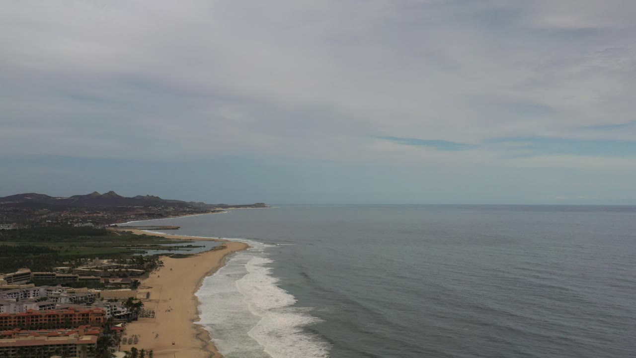 Aerial view of the Los Cabos coastline at the start of the clip, with gray-blue ocean, white surf, sandy beach, distant hills, shoreline buildings, and no visible people.