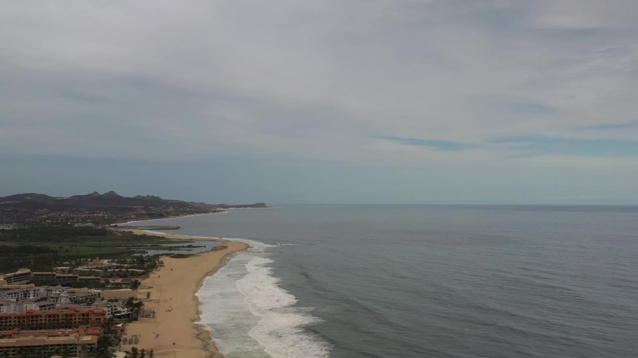 Wide aerial frame over Los Cabos showing waves curling onto the beach, open ocean to the right, low coastal buildings at lower left, distant hills, and no visible people.