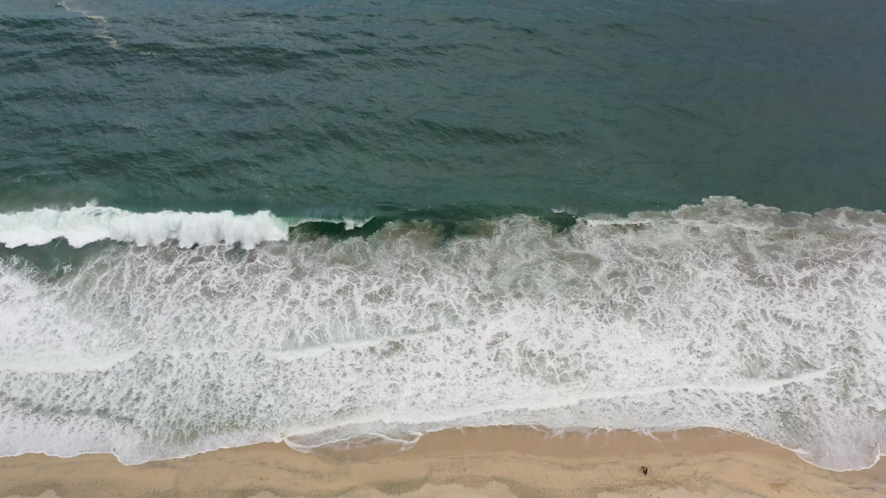 Overhead Los Cabos shoreline frame with a broad band of white foam pushing over wet sand and a small dark human figure near the lower beach.
