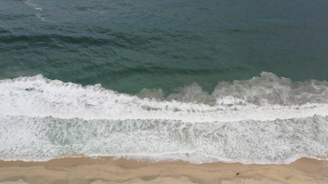 Overhead view in Los Cabos, Mexico, with dark teal ocean at the top, a foamy white wave crossing the middle, tan sand below, and no visible people.