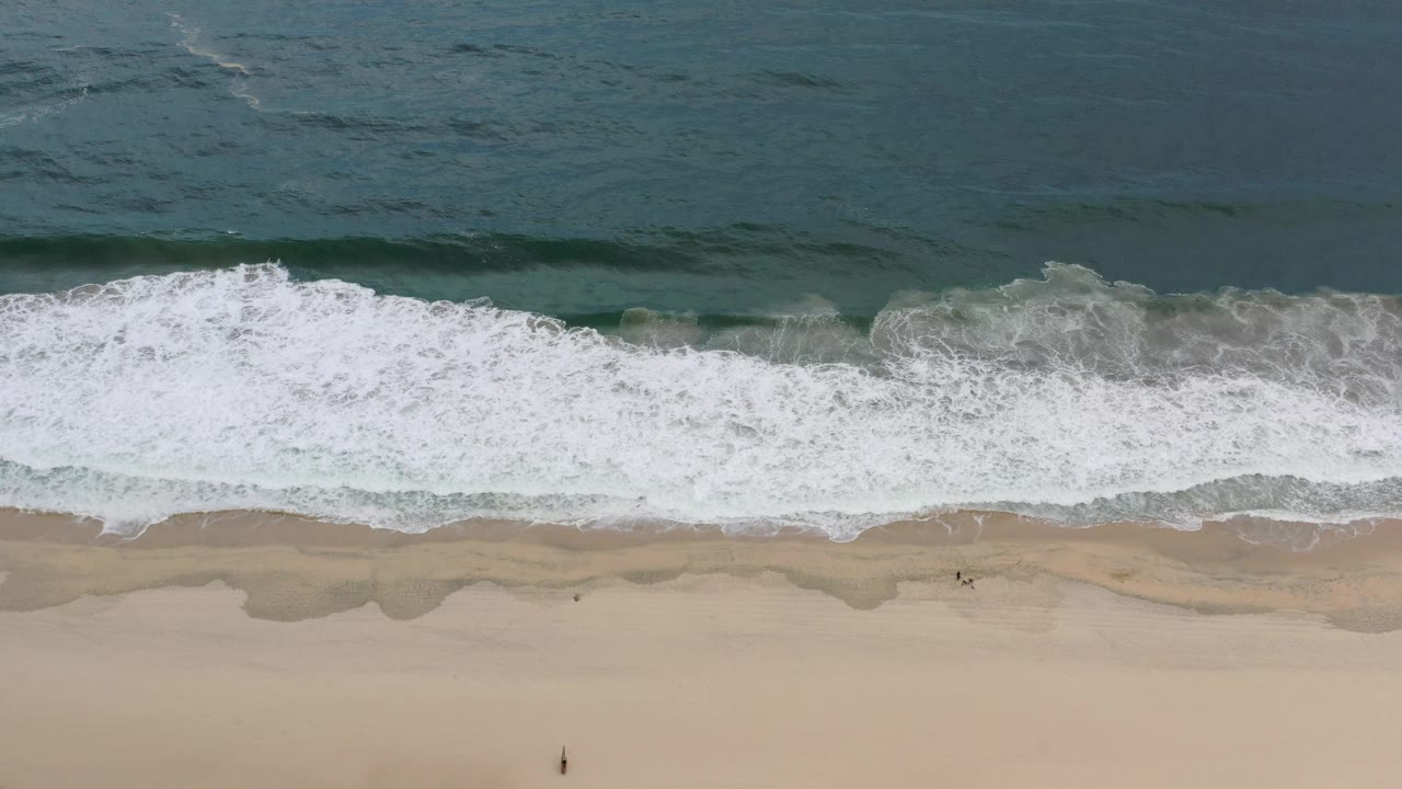 Overhead Los Cabos shoreline with a wider white foam band, darker water beyond it, wet sand scallops, and small people visible near the lower beach.