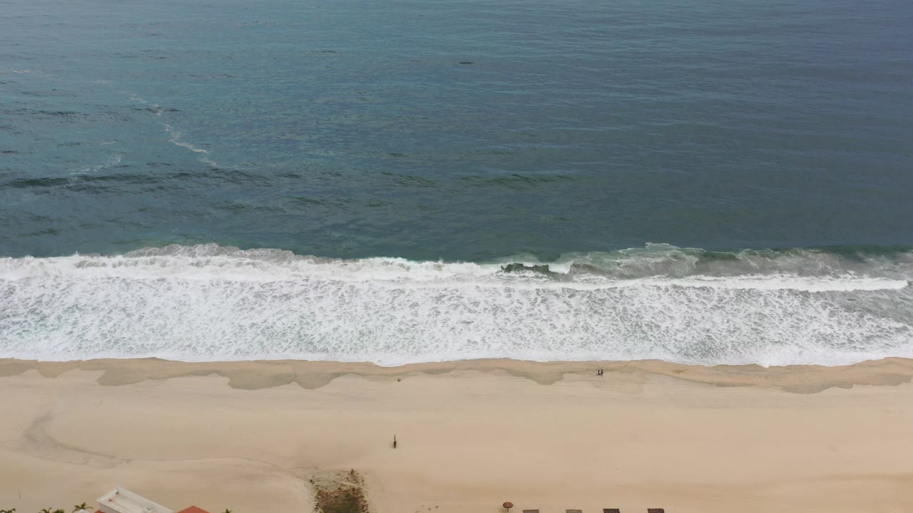 Aerial view in Los Cabos, Mexico, with blue-green water at the top, white surf breaking across the middle, pale sand below, and no visible people.