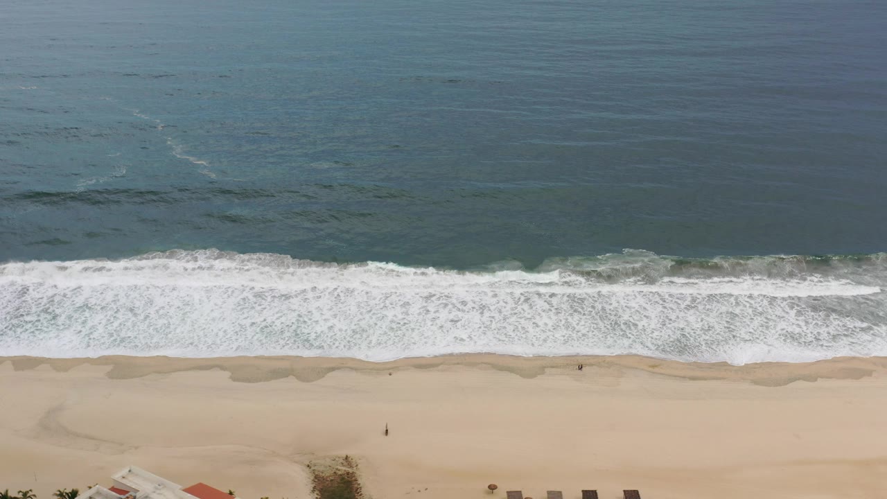Blue ocean and foamy breakers run parallel to an open sandy shore.