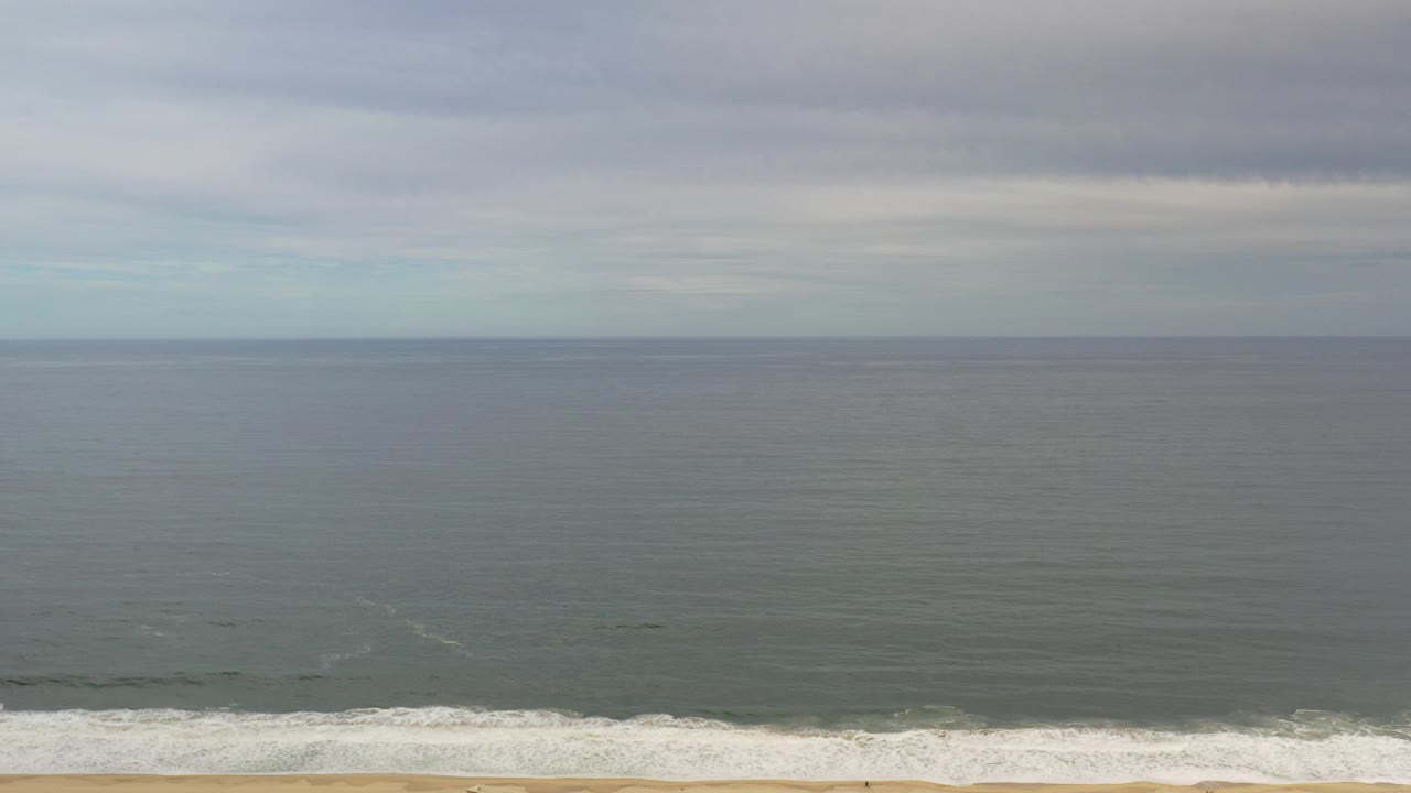 Wide ocean horizon under gray clouds with surf along a sandy beach.