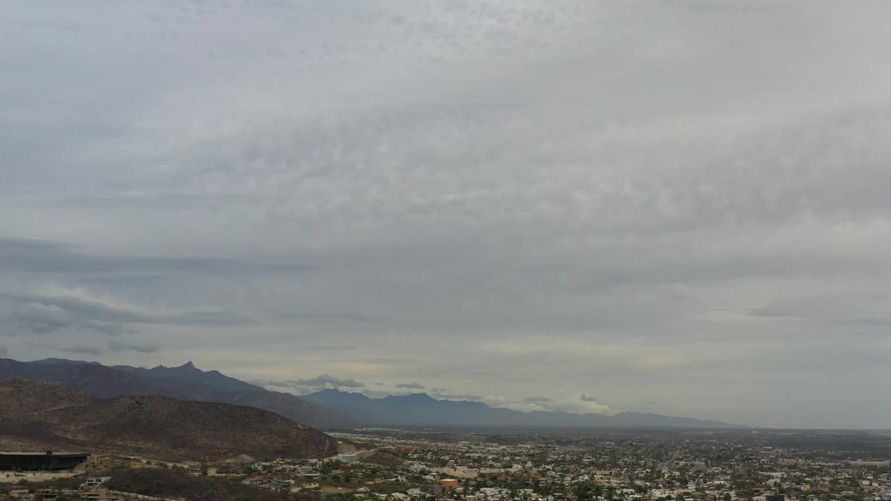 Los Cabos, Mexico, appears from a high viewpoint with city rooftops along the lower frame, dark hills to the left, distant mountains, layered gray clouds, and no visible people.