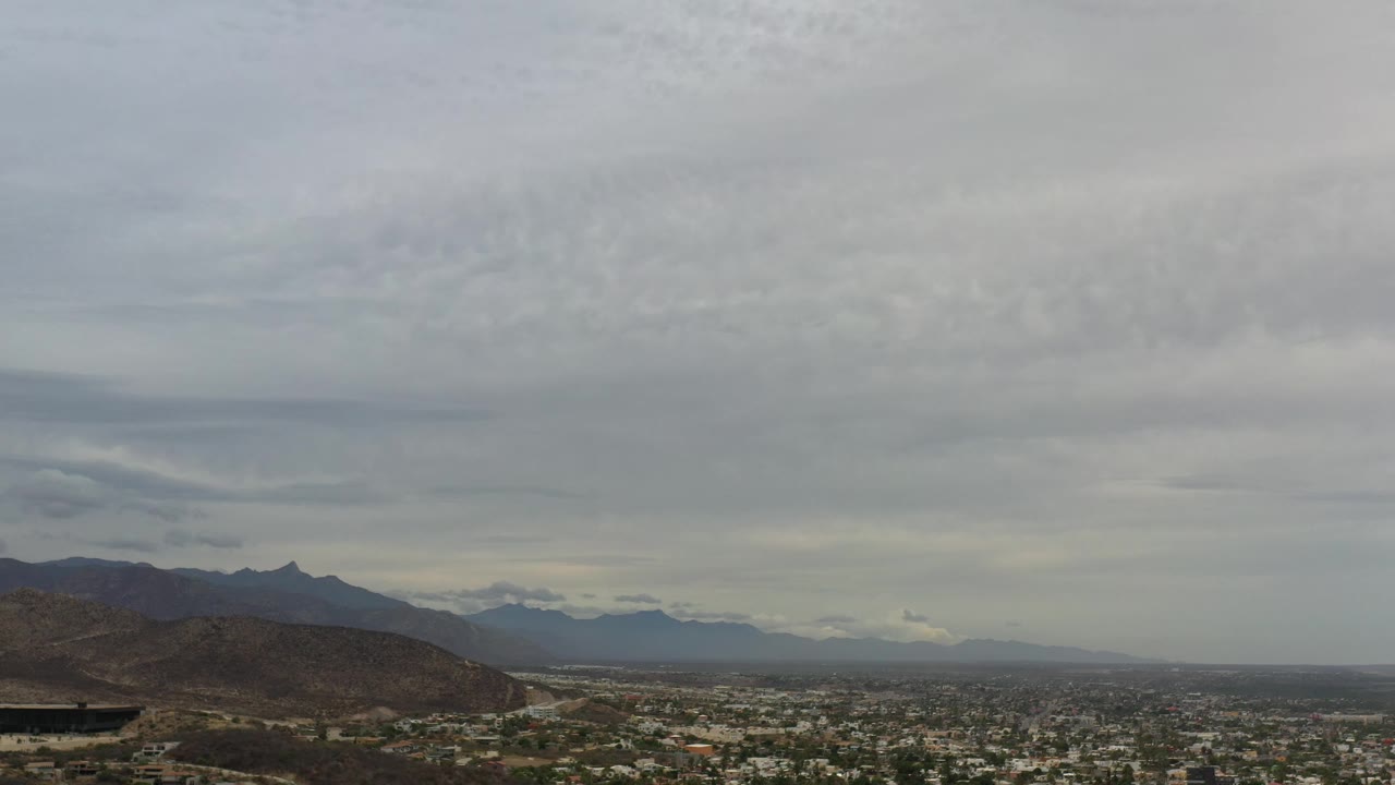 Los Cabos cityscape remains beneath layered gray clouds, with a dark mountain ridge on the left and hazy blue-gray landforms in the distance, no visible people.