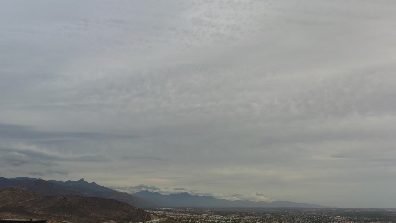 Opening frame of a Los Cabos landscape with dark hills at the lower left, hazy distant terrain, layered gray clouds, and no visible people.