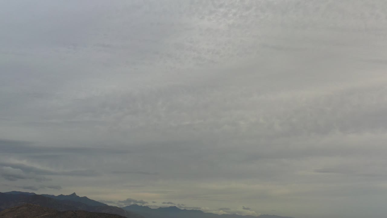 Los Cabos, Mexico view with light gray clouds filling the frame, a low mountain horizon, thin ground detail at the bottom, and no visible people.