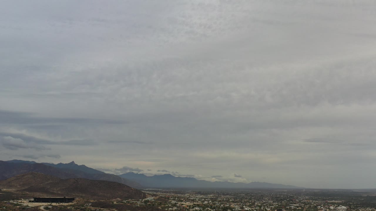 Wide aerial view of hills, city, distant mountains, and gray cloud cover.