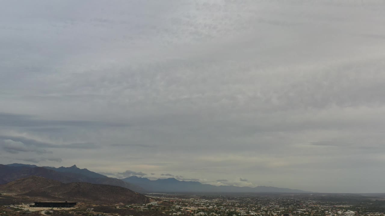 Los Cabos aerial view with a dark hillside in the lower left, city blocks stretching across the valley, hazy mountains beyond, and no visible people.