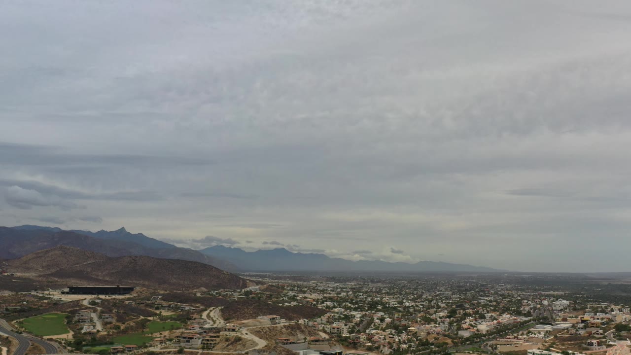Aerial panorama of Los Cabos showing houses and roads across the lower frame, a rounded hill at left, distant mountain ridges, and no visible people.