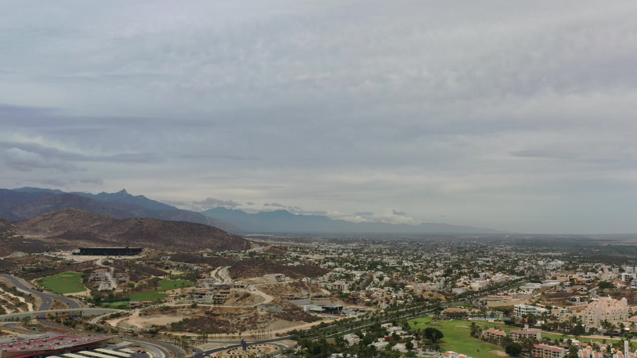 Wide aerial view of hills, roads, buildings, green fields, and gray clouds.