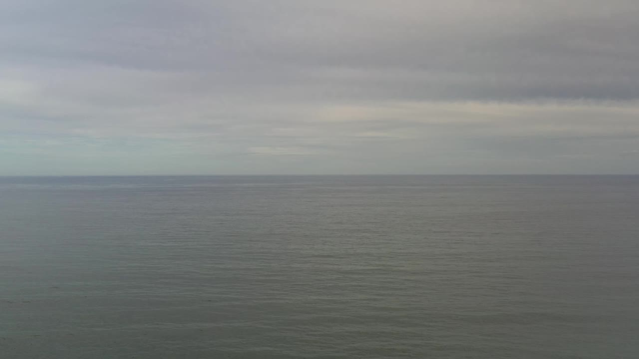 Gray ocean surface fills most of the frame with a thin horizon line and layered cloud cover above Los Cabos, Mexico, with no visible people.