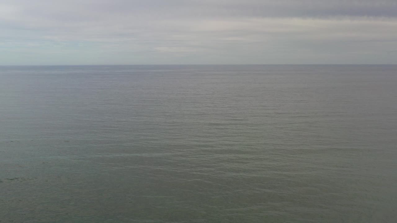Aerial view over the ocean in Los Cabos, Mexico, with a level horizon, gray cloud cover, muted blue-green water, and no visible people.