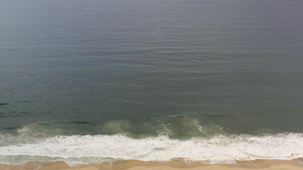 Overhead view of a Los Cabos, Mexico shoreline, with blue-green water above a white surf band and tan beach below, no visible people.