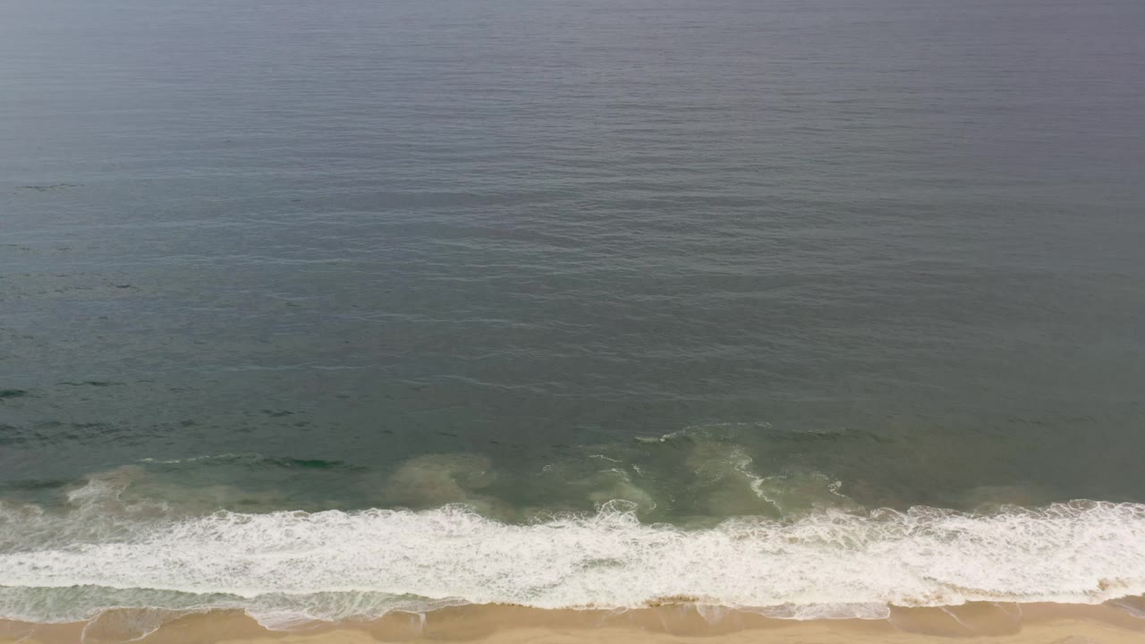 Aerial Los Cabos beach frame with the ocean occupying most of the image and a narrow strip of sand beneath the foamy wave line, no visible people.