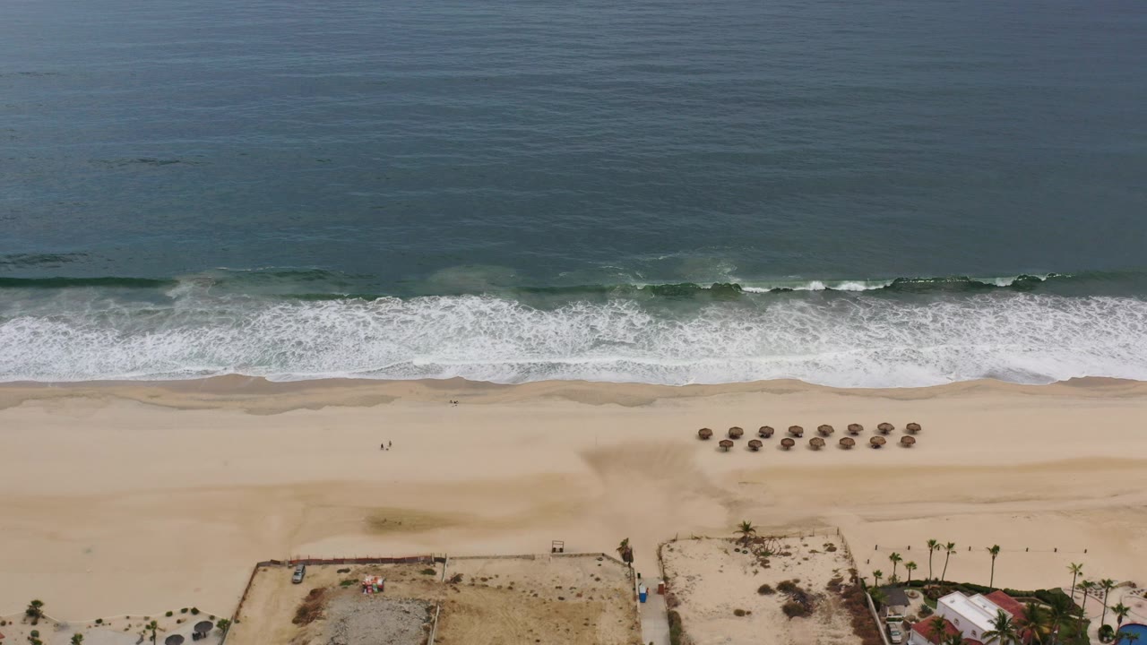 High aerial frame over a Los Cabos beach with teal water at top, white surf bands, pale sand, umbrella rows, and beachfront lots below, with no visible people.