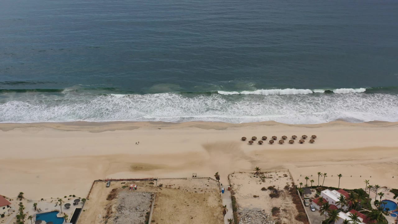 Drone view of surf, broad beach, umbrella rows, and beachfront homes with pools.