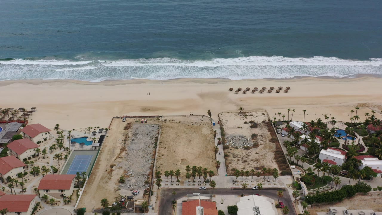 Aerial frame over a Los Cabos beach with blue ocean at top, white surf on pale sand, beachfront pools and red-roof buildings below, and no visible people.