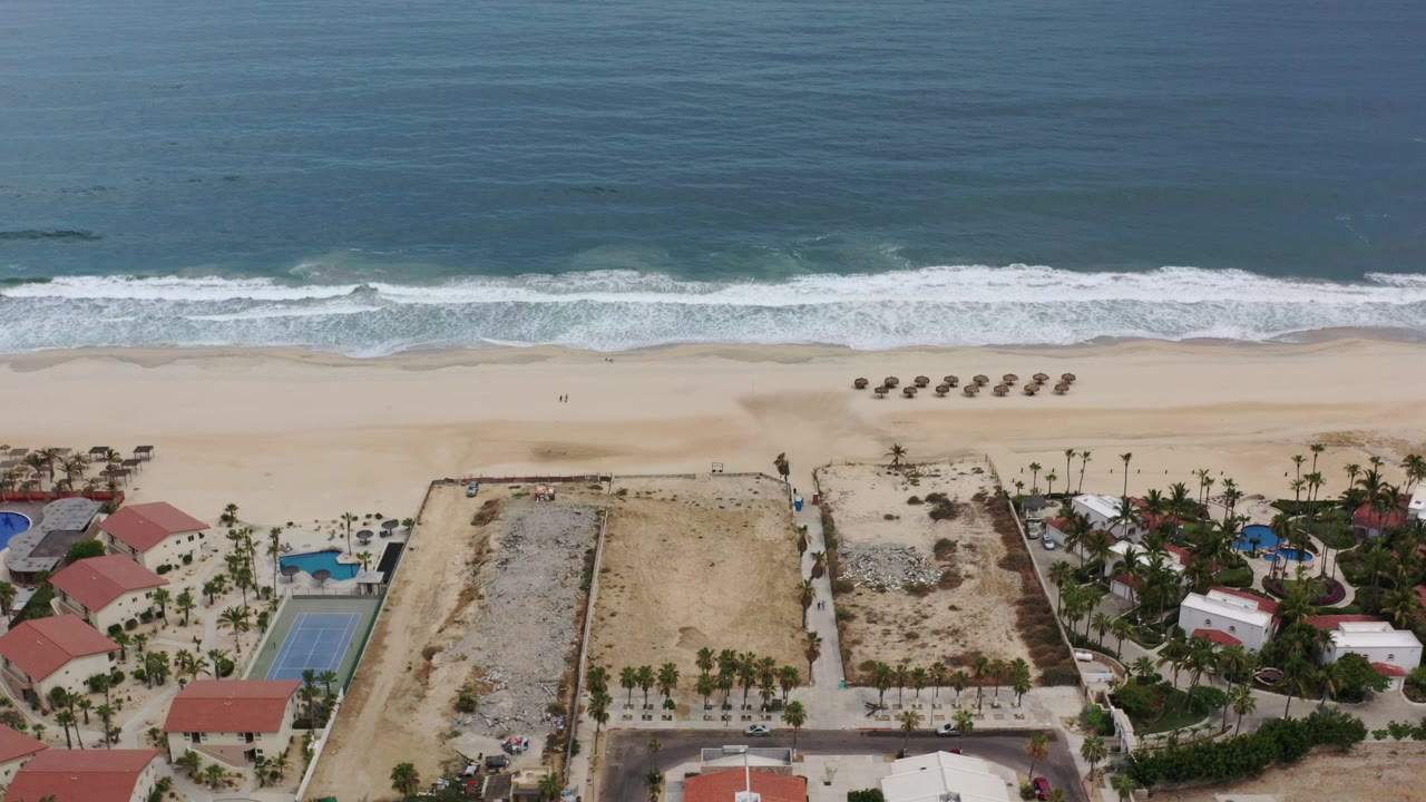 Overhead coastline with ocean, surf, beach umbrellas, palms, pools, and buildings.