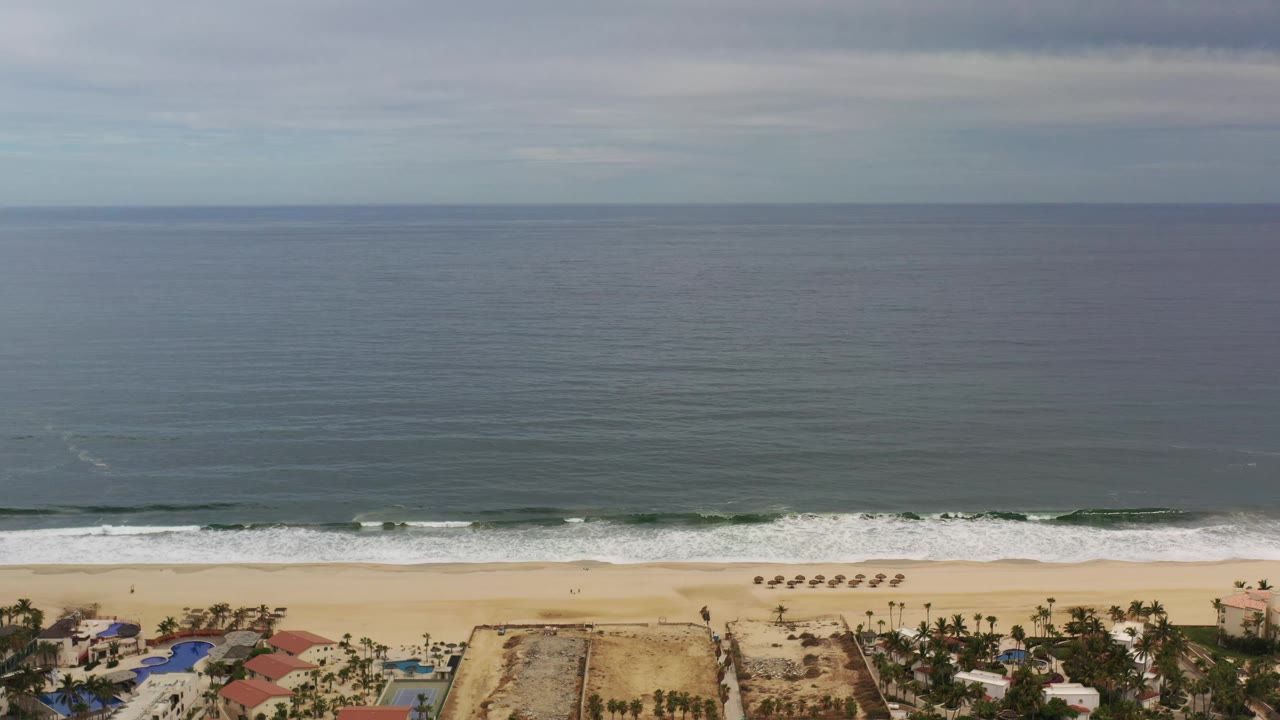 A Los Cabos aerial frame with the ocean occupying most of the view, a narrow white wave line at the shore, and small beachfront structures visible along the lower border with no visible people.