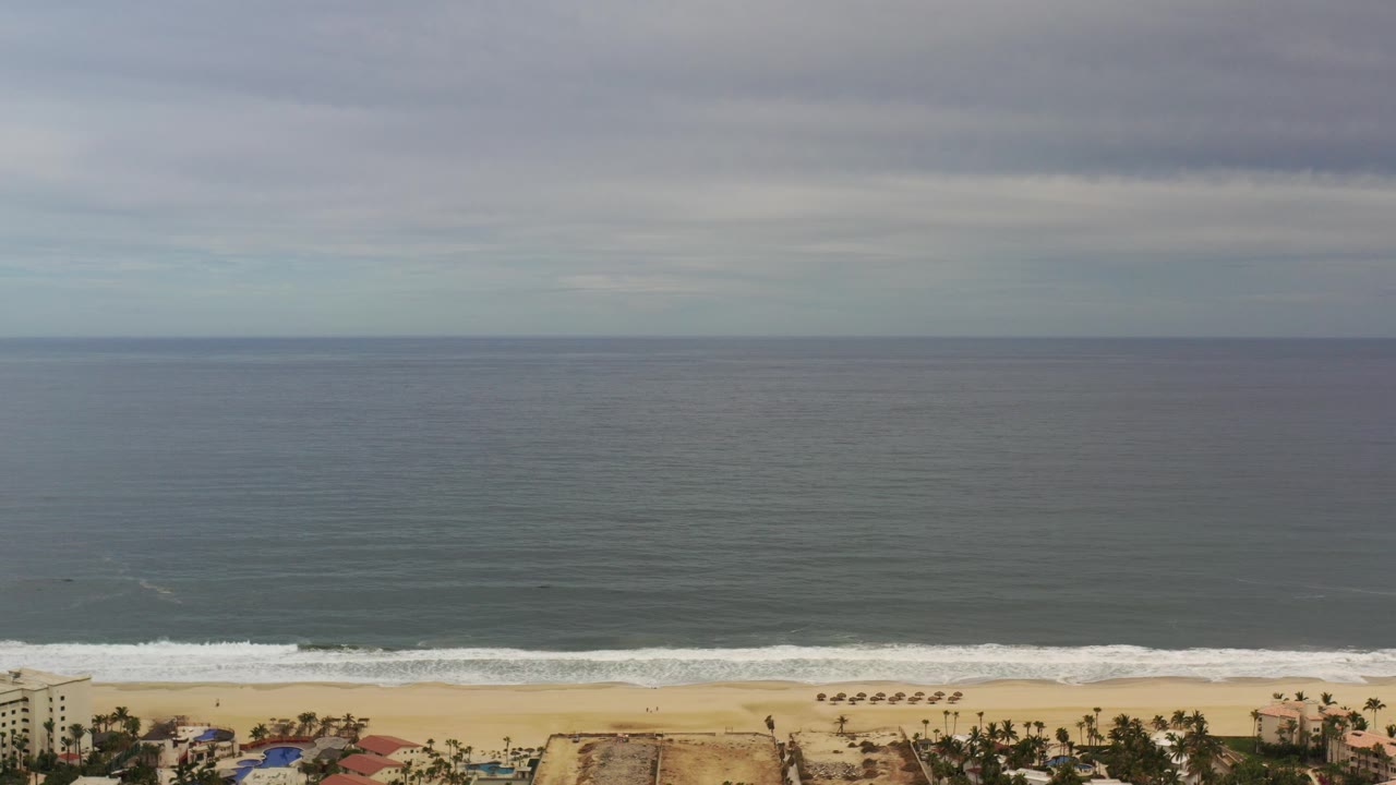Opening frame of Los Cabos ocean and beach from an elevated view, with dark water, gray cloud cover, a thin surf line, and no visible people.