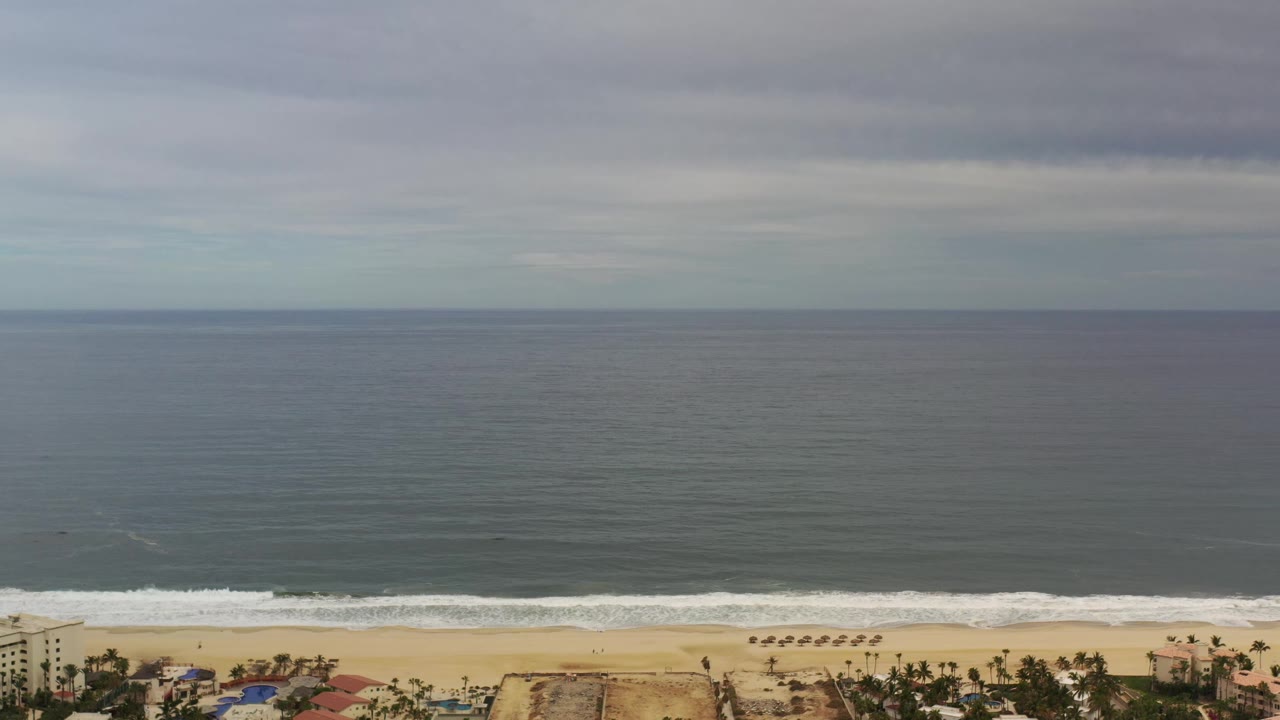 Second frame shows the Los Cabos shoreline held low in frame, with white waves rolling toward sand below a wide expanse of dark ocean and clouded sky, and no visible people.