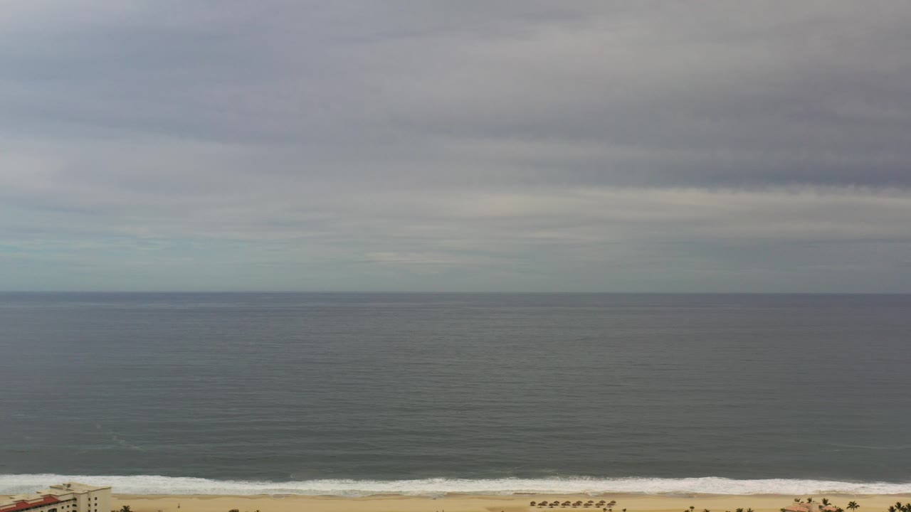 Wide Los Cabos beach view with gray water, layered clouds, a narrow strip of sand, and tiny people visible near the lower edge.