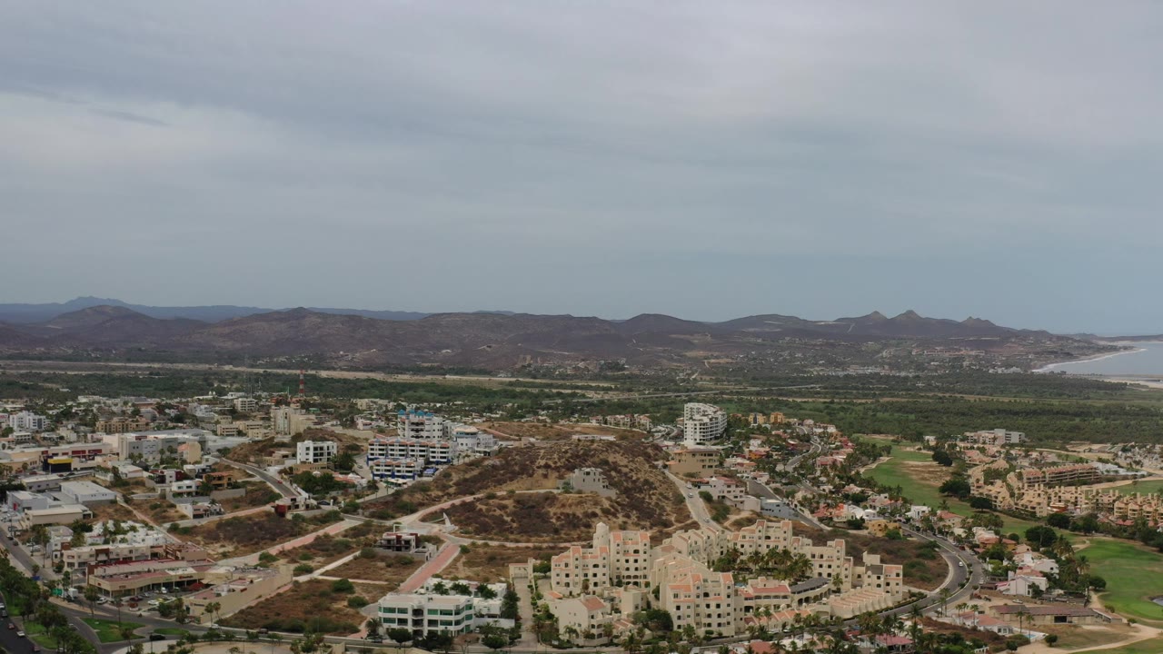 Wide aerial view of hillside buildings, roads, green areas, distant bay, and mountains under clouds.