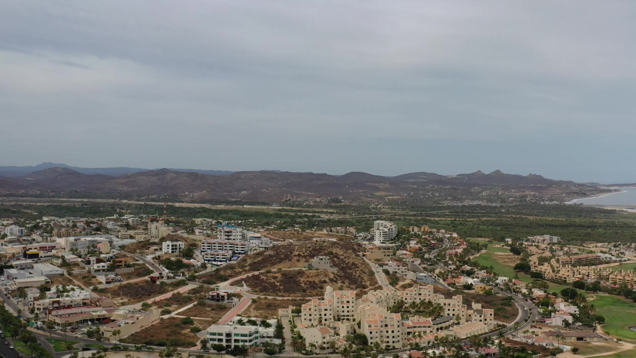 Elevated Los Cabos, Mexico frame with clustered buildings, curving roads, brown hills, green strips, a pale shoreline in the distance, and no visible people.
