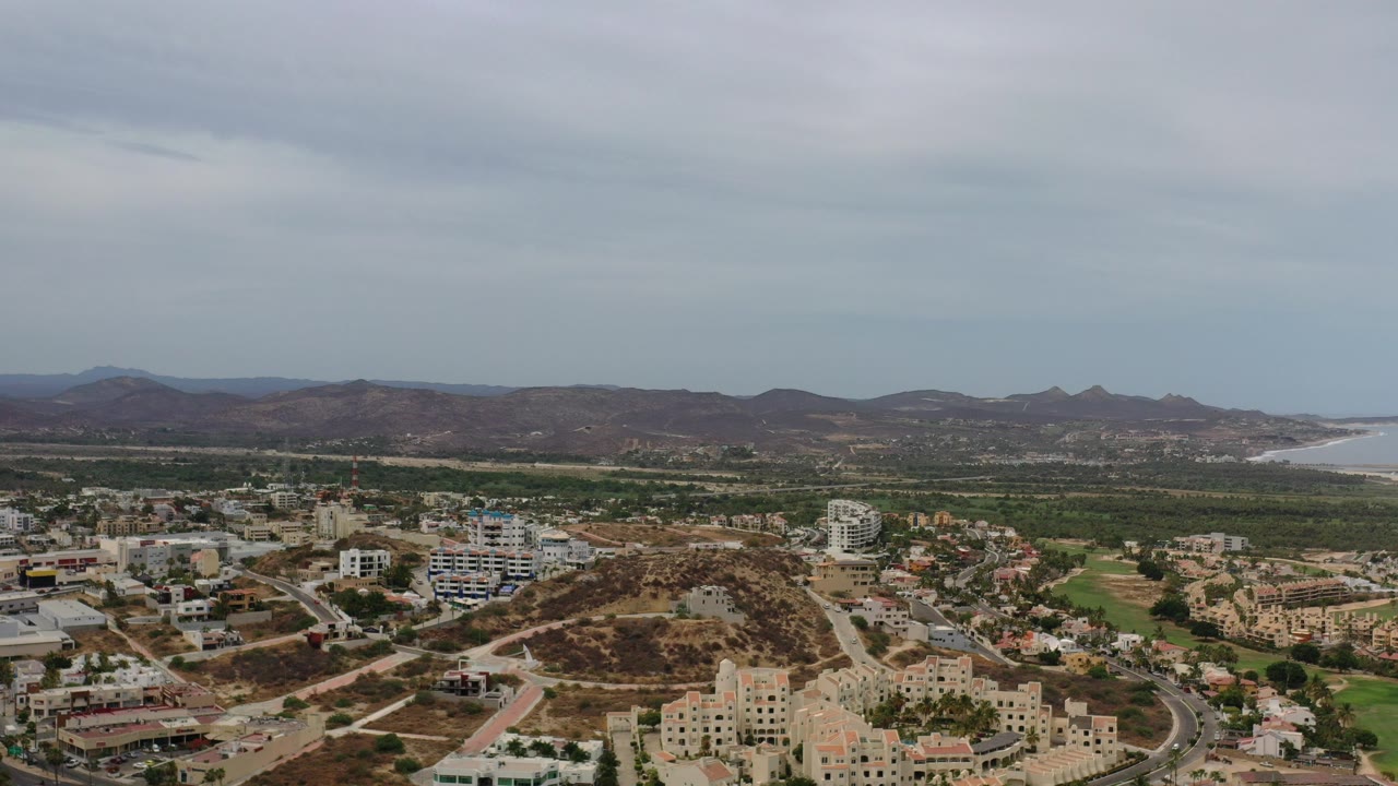 Wide aerial view of buildings, roads, desert hills, distant coast, and cloudy sky.