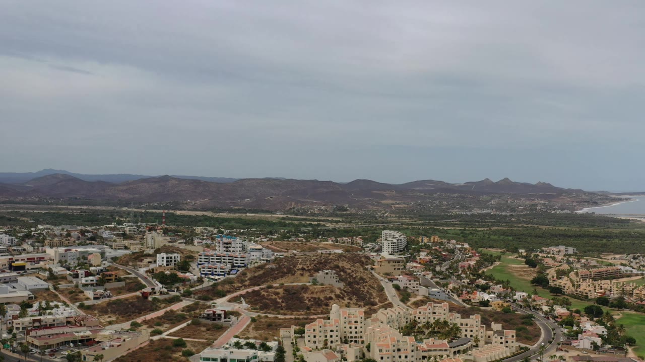 Early aerial frame over Los Cabos, Mexico, with a brown hillside and residential blocks in front, green vegetation through the middle distance, and no visible people.