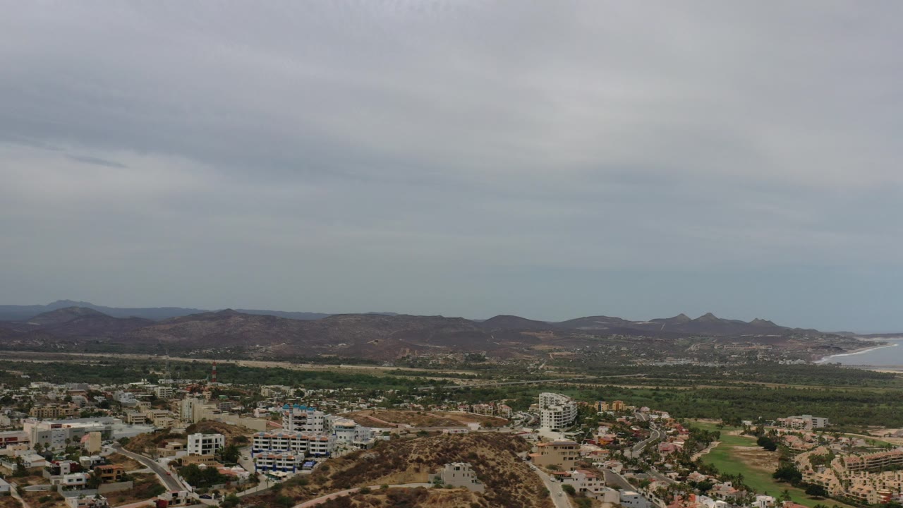 Wide aerial frame over Los Cabos, Mexico, showing low buildings in the foreground, green fields, brown hills, a distant bay, and no visible people.
