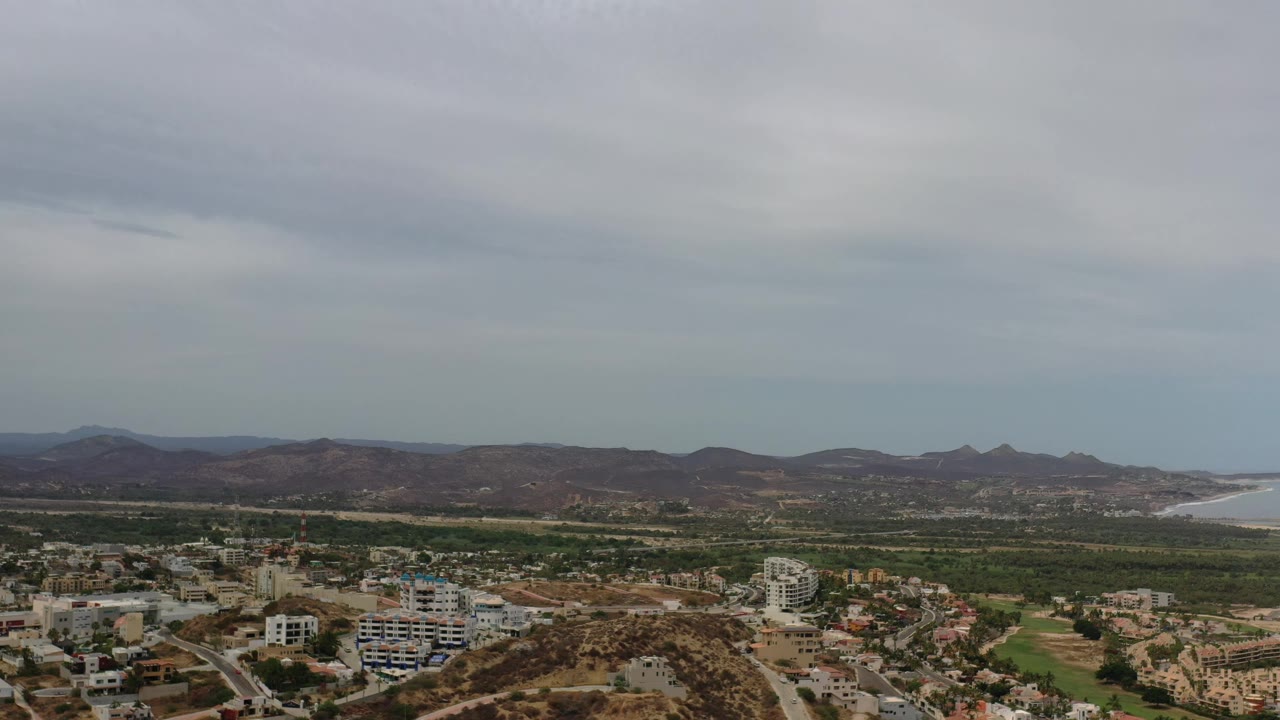 A high Los Cabos aerial view shows clustered buildings at the bottom of frame, a band of green land, rugged hills, a pale bay, and no visible people.