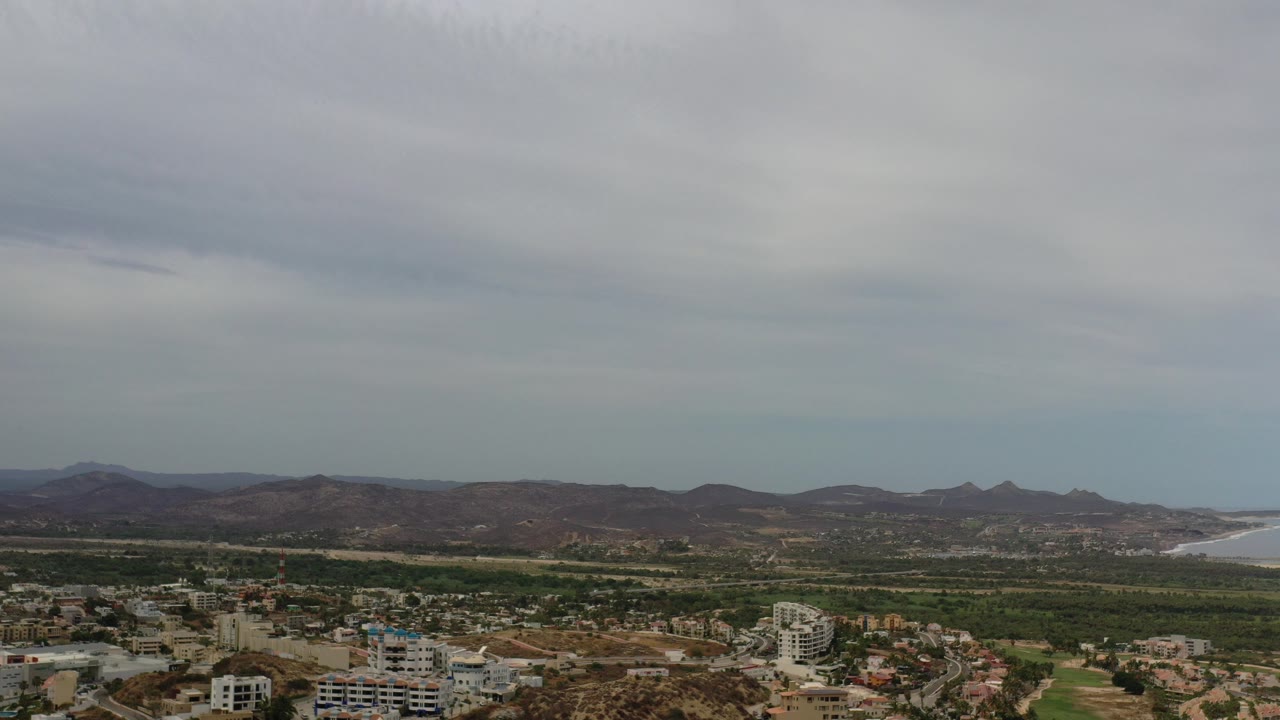 Opening frame of a Los Cabos aerial view with low-rise buildings below, a green band of land across the middle, distant hills, ocean at the right edge, and no visible people.