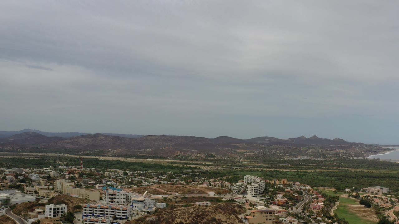 Opening Los Cabos aerial view with hillside buildings in the foreground, green lowland areas, distant brown mountains, a shoreline at the right edge, and no visible people.