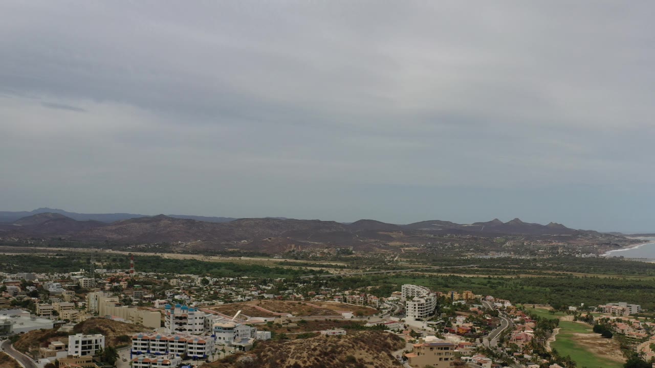 Wide Los Cabos aerial frame with white and tan buildings in the lower frame, green terrain across the center, brown mountain ridges behind it, a pale beach line at right, and no visible people.