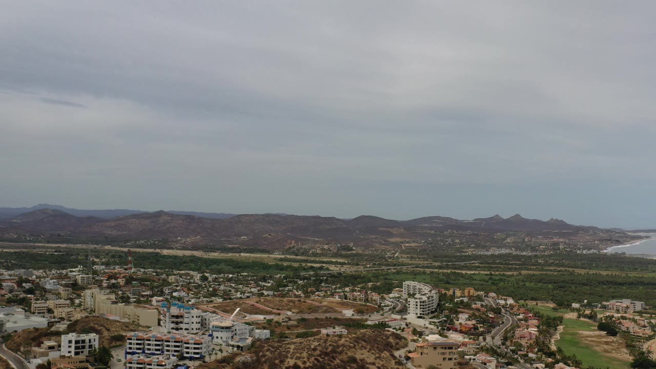 Broad elevated view of rooftops, roads, green land, mountains, and distant shoreline beneath clouds.