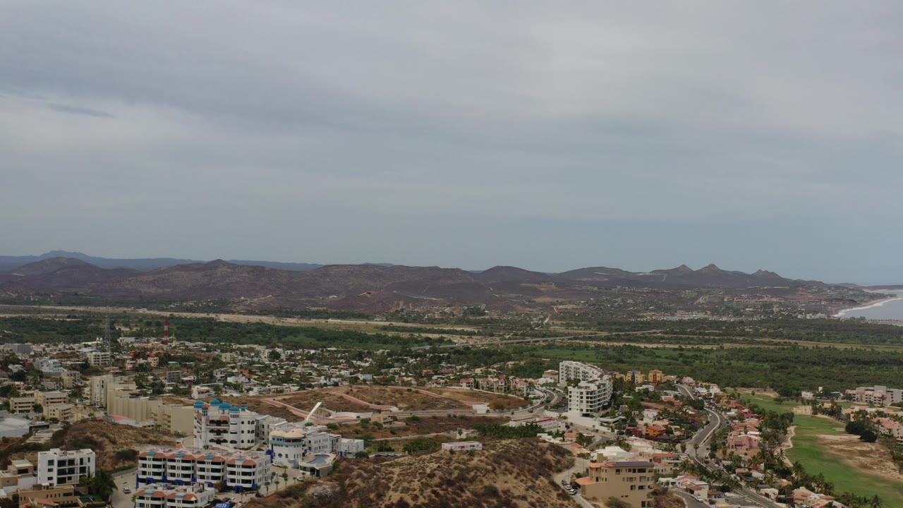 Opening aerial frame over Los Cabos with residential buildings in the foreground, green landscape through the center, brown mountains on the horizon, distant coastline, and no visible people.